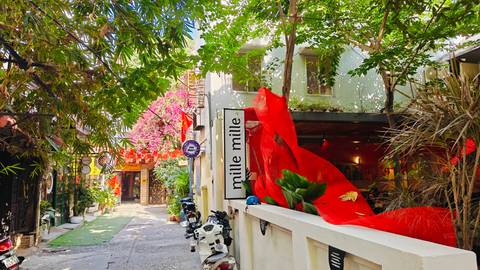 Colorful alley with bougainvillea, a cafe sign reading 'mille mille', scooters, and vibrant red decorations under bright daylight.