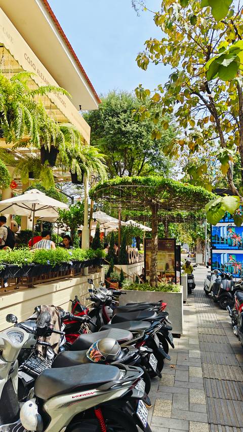 Outdoor street-side cafe with umbrellas, lush plants, and patrons enjoying drinks in a leafy urban setting.