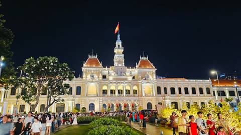 Illuminated French-colonial style city hall with Vietnamese flag at night, crowds gathered in landscaped plaza.