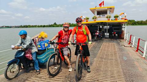 Cyclists with mountain bikes on a small river ferry, another passenger on scooter beside them under bright skies.