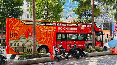 Bright red double-decker hop-on hop-off tour bus labeled 'Ho Chi Minh' parked under palm trees.
