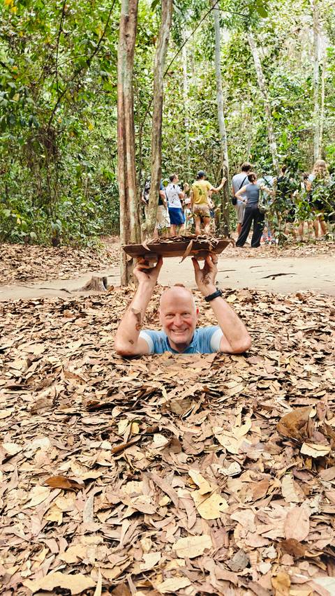 Smiling tourist emerging vertically from a narrow jungle tunnel camouflaged with dry leaves at Cu Chi.