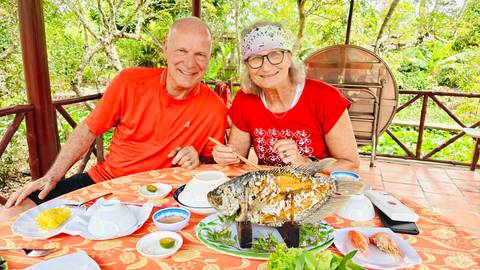 Smiling couple at garden restaurant table with whole grilled fish and Vietnamese dishes laid out.