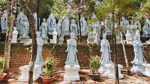 Terraced rows of white stone Quan Âm statues set against a brick retaining wall and dappled forest light.