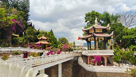 Multi-level pagoda complex with ornate bridges, bougainvillea, and mountainous backdrop under partly cloudy sky.
