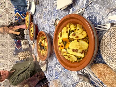 Traditional Moroccan tagine dishes served on patterned blue table with diners around.