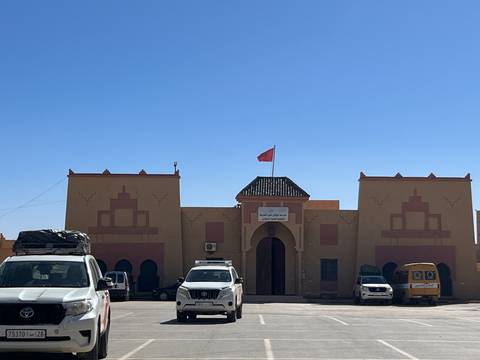 An adobe-style Moroccan public building with a flag and parked vehicles under a clear sky.
