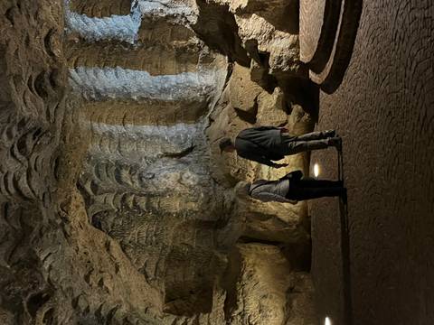 Two visitors observe textured rock formations inside a dimly lit cave chamber.