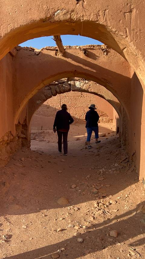 Two women walking through an arched, earthen ruin passageway with sunlit desert backdrop, likely in Ait Benhaddou area