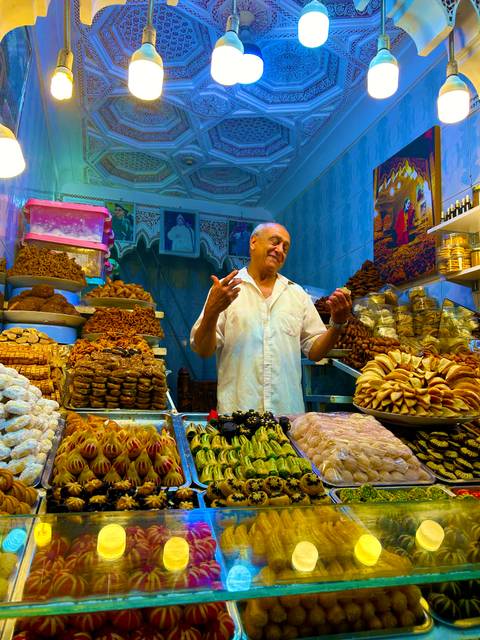 Smiling Moroccan sweets vendor standing behind colorful display of pastries inside a medina shop