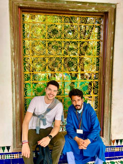 Two men sitting by ornate golden lattice window frame in Moroccan building, smiling at camera