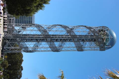 A tall, modern metal tower with a spiral lattice design and a glass sphere on top, set against a clear blue sky.