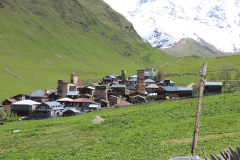A mountain village with traditional stone defensive towers nestled on a green hillside, with snow-capped peaks in the background.