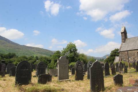 Old hillside cemetery with stone headstones beneath partly cloudy blue sky and rolling green mountains.