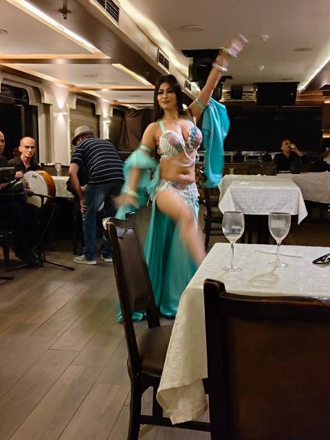 Belly dancer in turquoise costume performing between dining tables with live musicians nearby