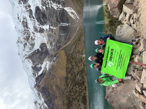 Trekking team holds bright green Machu Picchu Expeditions banner beside emerald Humantay Lake and snowy peaks