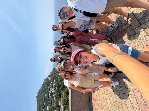 A cheerful group of young travelers taking a wide-angle selfie on a sunny terrace with coastal cliffs, hillside homes and the deep-blue sea behind them.