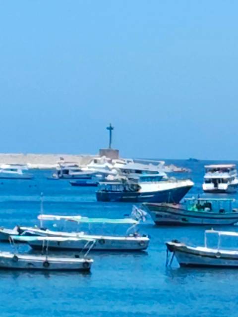 Blurry view of small boats and yachts on bright blue water near a low harbour wall and a tiny lighthouse