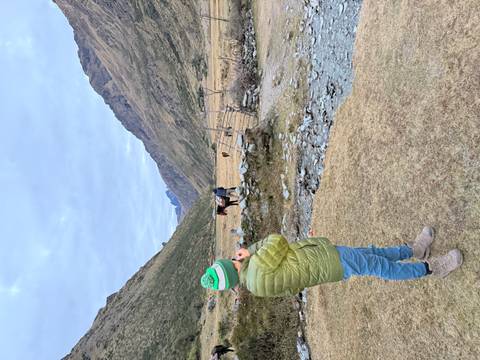 A youngster in a green jacket watches a companion ready horses in a broad mountain valley under an overcast sky.