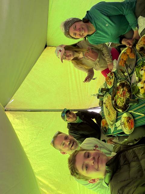 Three trekkers enjoy a colourful meal laid out on a striped camping table inside a tent.