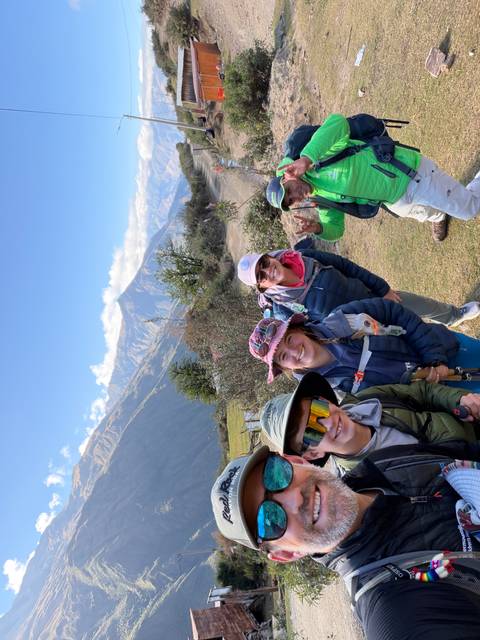 Four happy trekkers pose for a selfie with sun-lit Andean peaks and blue sky in the background.