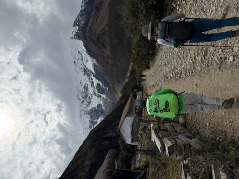 Trekkers walk a stony path toward a snow-capped peak with tents and low clouds ahead.