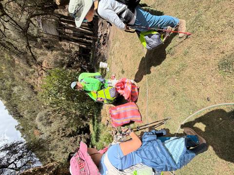 Porters rest at a grassy camp area with colorful sacks and gear under dappled sunlight.