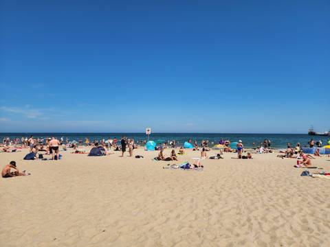 Crowds sunbathe and play on a wide sandy Baltic beach beside deep blue water under a clear sky.