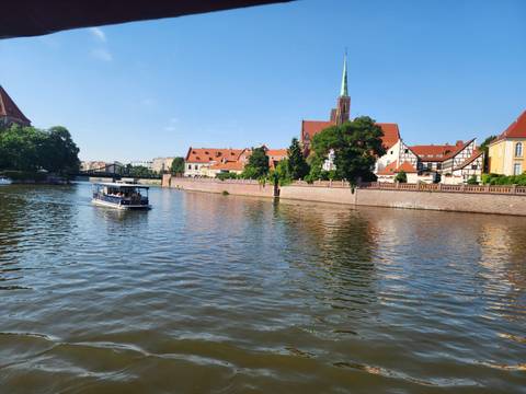 Tour boat cruises a calm river past red-roofed churches and leafy embankments on a bright day.