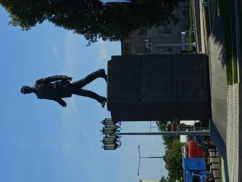 Bronze statue of a walking figure atop a tall dark plinth set against a clear blue sky.