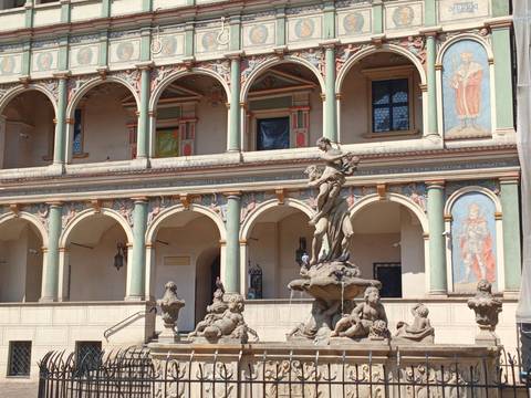 Renaissance town hall facade with colorful frescoed arches and an ornate fountain in front under bright sunlight.
