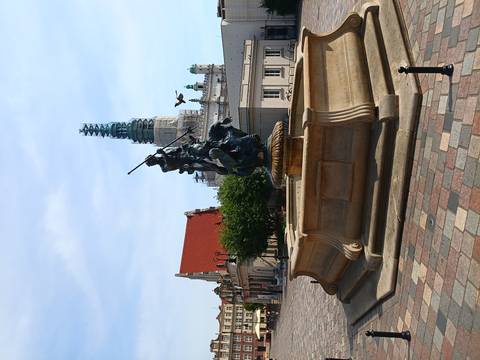 Stone fountain featuring a mounted knight statue on a historic market square with a scaffold-covered tower behind.