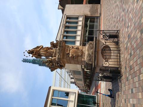 Baroque column monument topped by a crowned saint surrounded by stars on a paved plaza beside modern buildings.