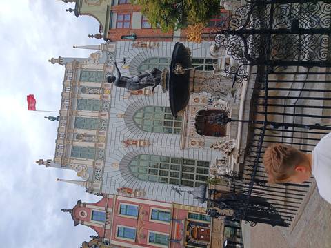 Neptune Fountain in front of the ornate Artus Court building flying the Polish flag, with a child observing in the foreground.