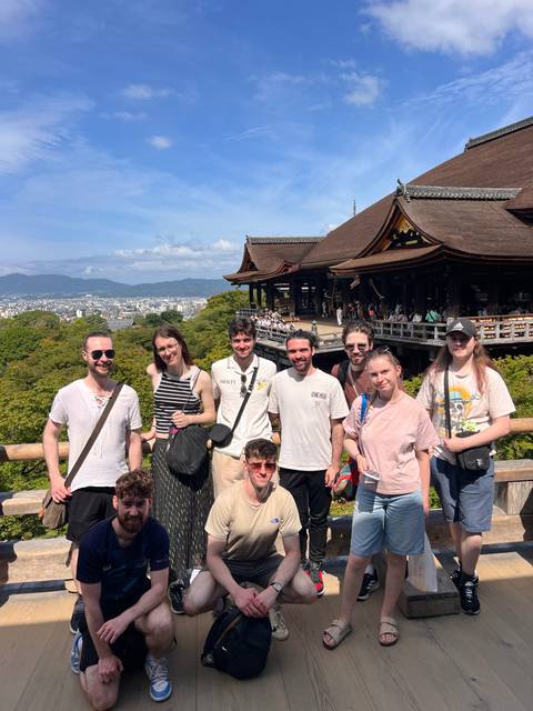 Tour group poses with city panorama and wooden veranda of Kiyomizu-dera temple behind them.