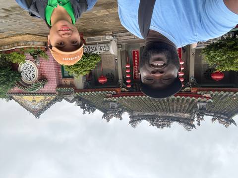 Two travelers pose in front of an ornate temple with green tiled roof and red lanterns on a cloudy day.