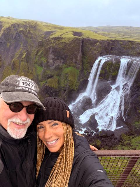 Smiling couple taking a selfie with a tall moss-covered waterfall cascading behind them