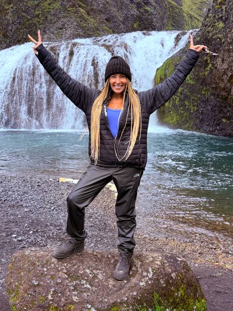 Woman in outdoor gear raising her arms joyfully beside a small waterfall pool