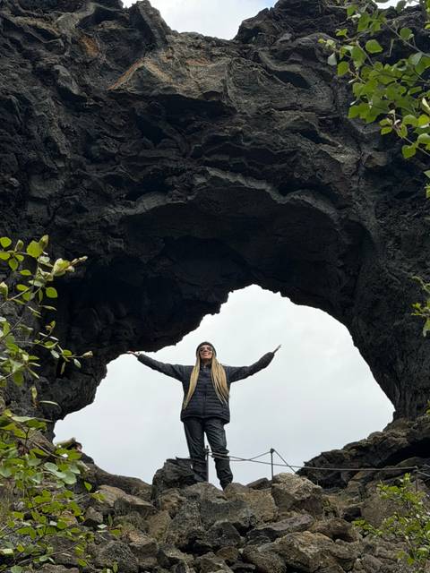 Woman standing triumphantly under a large natural lava arch against a cloudy sky