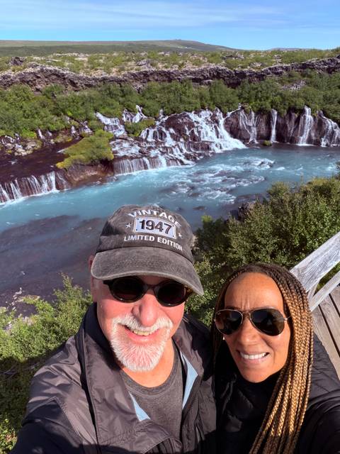 Selfie overlooking a wide blue cascade flowing through dark lava fields and shrubs