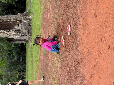 Small girl in bright clothes playing with dirt on a red clay ground in a forest clearing
