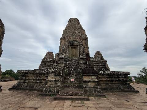 Tall stone temple tower on a platform against a cloudy sky, visitor seen in the distance