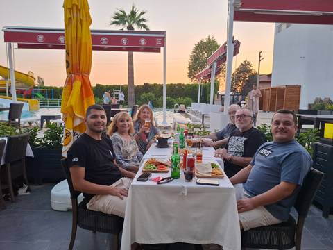 Six travelers enjoy dinner at a poolside restaurant, raising drinks around a table of local dishes.