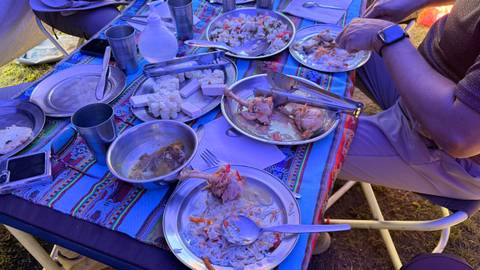 Camping meal on patterned cloth with partially eaten plates of food and two travelers seated around table.
