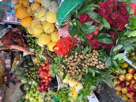 Vibrant fruit stall overflowing with tropical produce such as longan, melon and rambutan while a vendor selects items in the background.