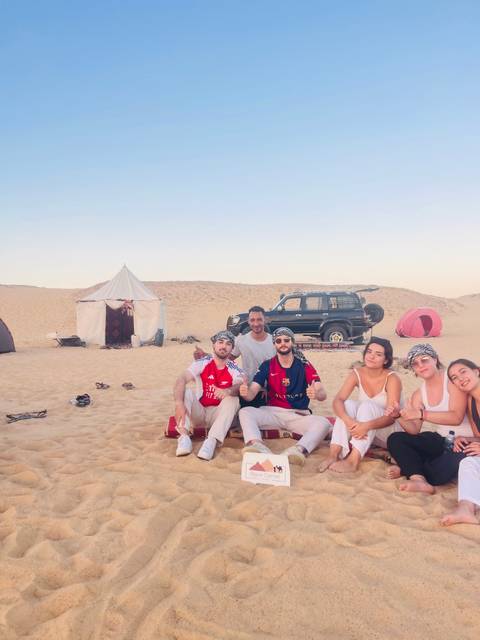 Group of friends relax on desert sand near tents and a 4x4 jeep during a Sahara camp.