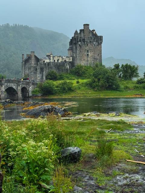 Iconic Eilean Donan Castle and stone bridge reflected in tidal waters with misty highlands behind.