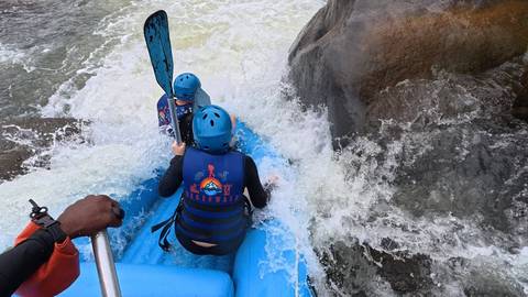 Adventurers in blue helmets and life-jackets paddle a raft through a churning rocky rapid.