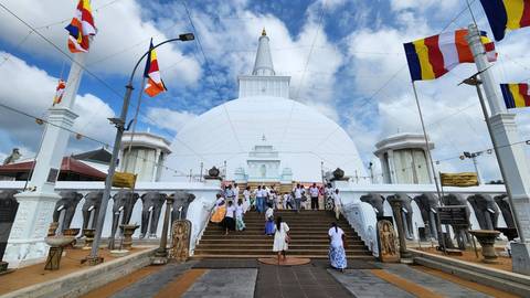Pilgrims in colorful dress ascend steps toward the vast white Ruwanwelisaya stupa under blue skies.