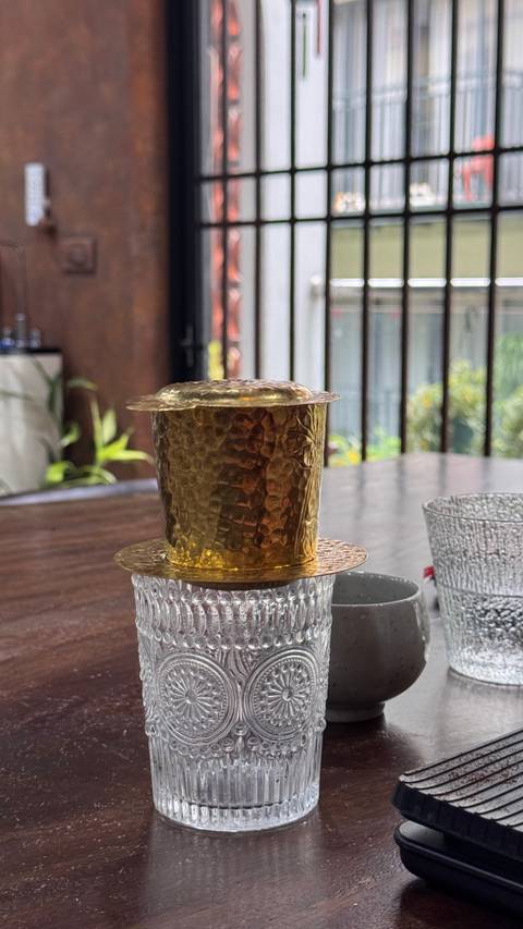 Close-up of a hammered brass Vietnamese coffee filter resting on textured glass cups indoors